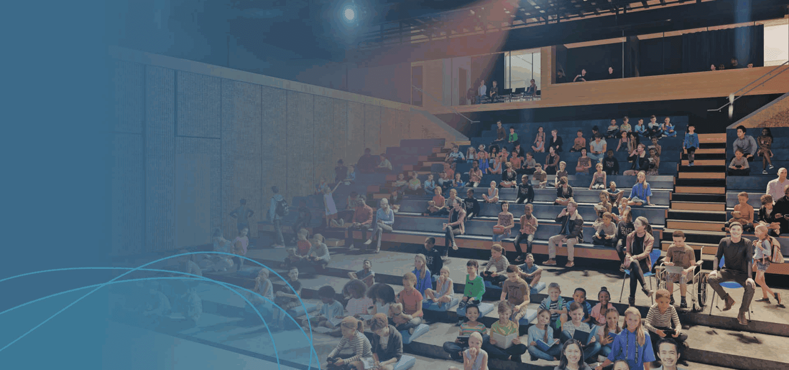 A large group of people, including children and adults, are seated on tiered benches in an indoor auditorium, facing the front.