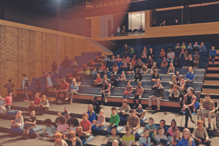 Children sit on tiered benches in a modern auditorium, some reading books, while others talk or look around.