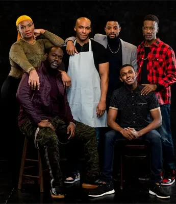 Six people pose together against a black background, with four standing and two seated on stools, all looking at the camera.