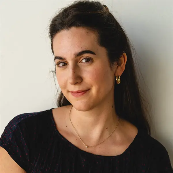 Woman with long dark hair, wearing a black top, gold hoop earrings, and a delicate necklace, standing against a plain light background and looking at the camera with a slight smile.