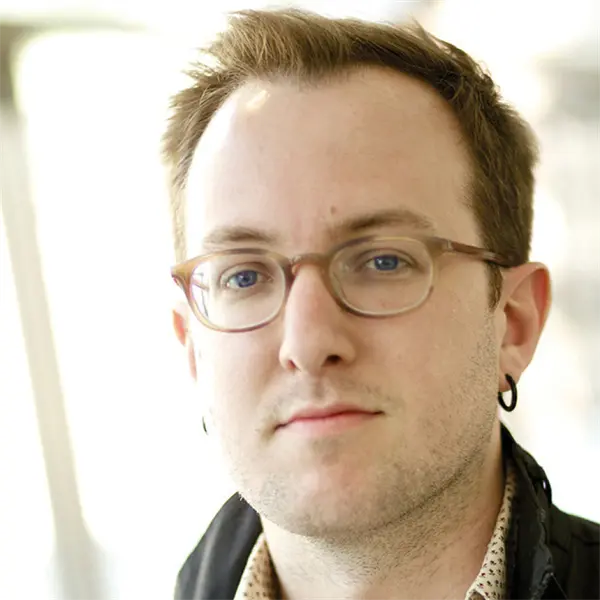 A man with short brown hair, glasses, and hoop earrings looks at the camera with a neutral expression. The background is softly blurred.