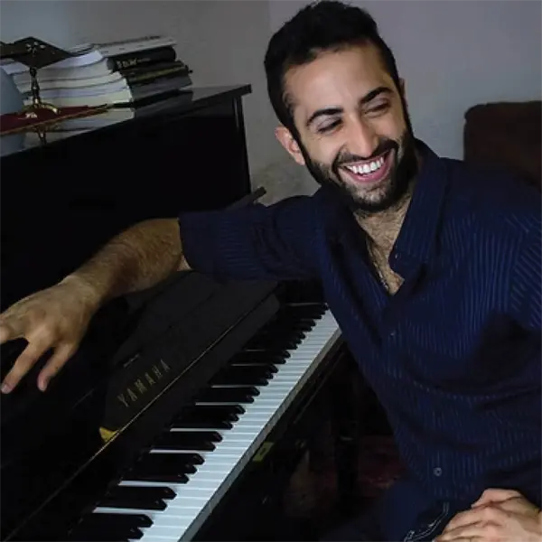 A man with a beard smiles while sitting at a Yamaha piano, resting one hand on the keys and the other on the piano lid.