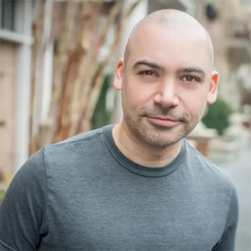 A man with a shaved head and a trimmed beard wearing a grey t-shirt stands outdoors, looking at the camera with a neutral expression.