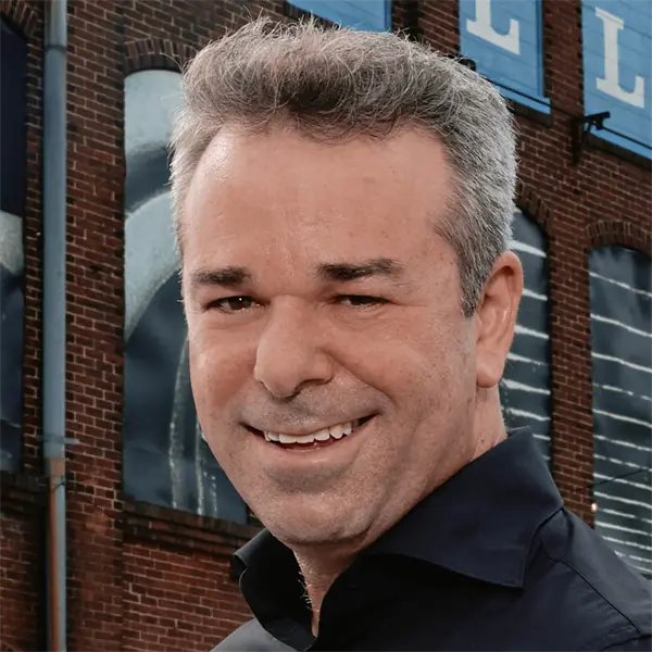 A man with short gray hair and a dark shirt smiles in front of a brick building with large blue and white signs.