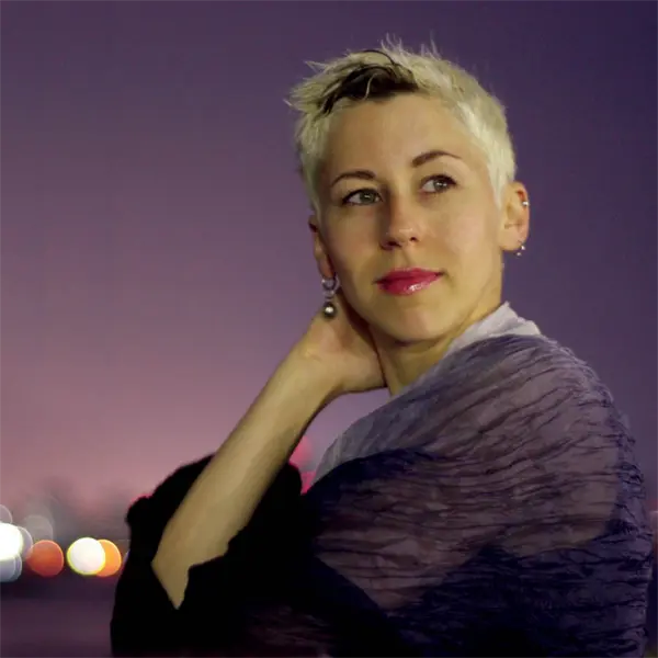 A person with short, blond hair looks to the side at night, with blurred city lights in the background.