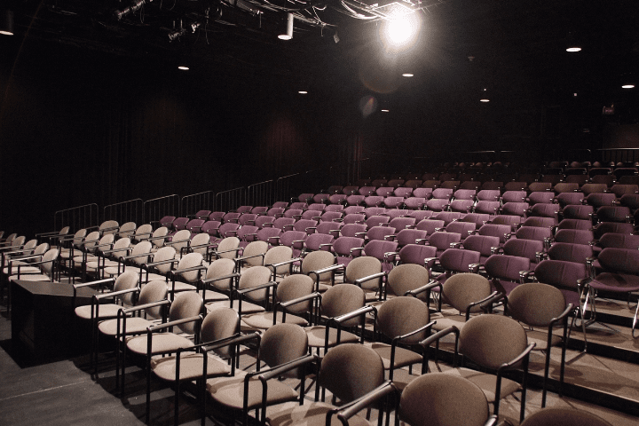 Rows of empty, purple and brown chairs face a stage in a dimly lit, modern theater auditorium.