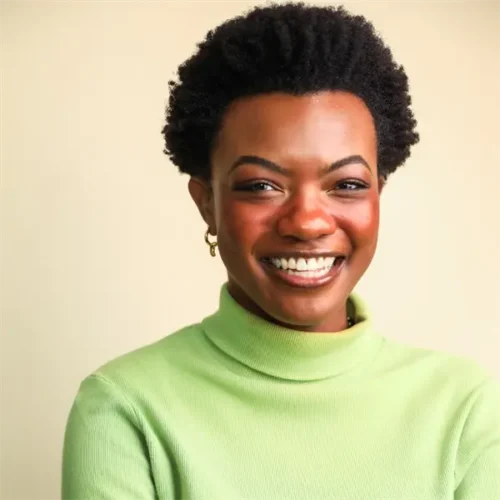 A woman with short curly hair, wearing a light green turtleneck, smiles while standing in front of a plain beige background.