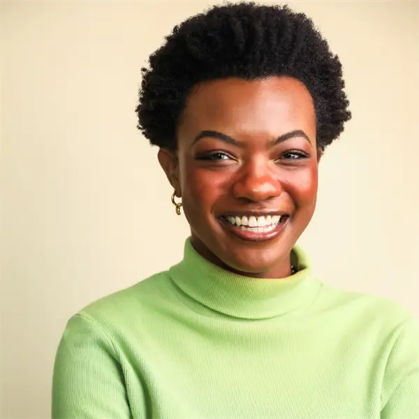 A woman with short curly hair, wearing a light green turtleneck, smiles while standing in front of a plain beige background.