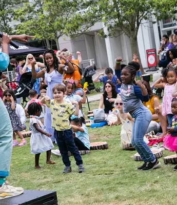 A woman with a microphone leads a group of children dancing on grass in an outdoor public space, with adults and more children watching in the background.