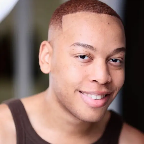 A person with closely cropped reddish hair and a brown tank top looks at the camera and smiles.
