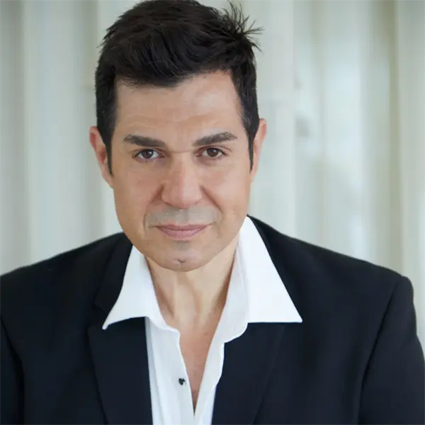 A man with short dark hair wearing a black suit jacket and white dress shirt stands in front of white curtains, looking at the camera.