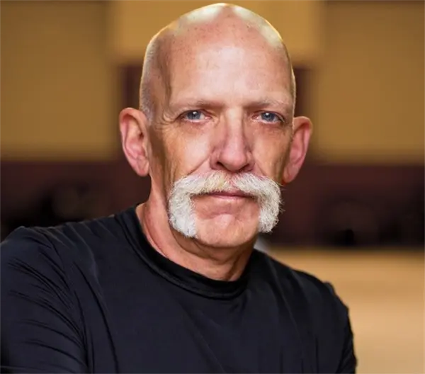 A bald man with a large white handlebar mustache wearing a black shirt stands indoors, looking directly at the camera.