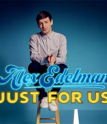 A man in a light-colored shirt sits on a stool against a blue background with the neon-style text "Alex Edelman JUST FOR US" displayed in front of him.