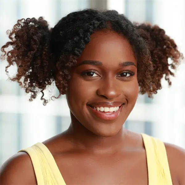 A woman with curly hair styled in two puffs smiles at the camera. She is wearing a yellow sleeveless top and is in front of a blurred background.