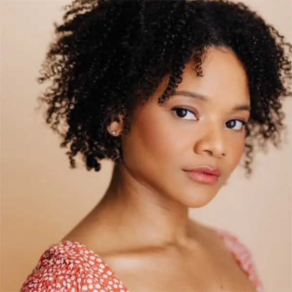 A woman with short curly hair and a red patterned top looks at the camera with a neutral expression against a beige background.