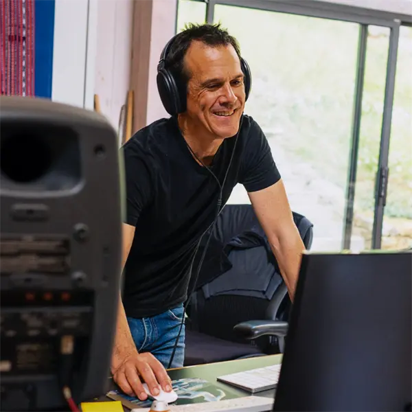 A person wearing headphones stands at a desk with audio equipment and a computer monitor, appearing to work in a music studio.