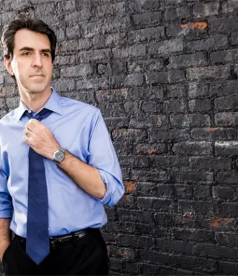 A man in a blue dress shirt and tie stands in front of a brick wall, adjusting his tie with one hand and looking to the side.