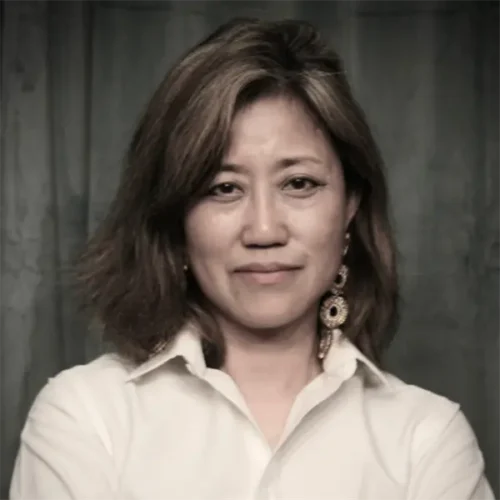 A woman with shoulder-length hair, wearing a white collared shirt and large earrings, stands in front of a dark background with her arms crossed.