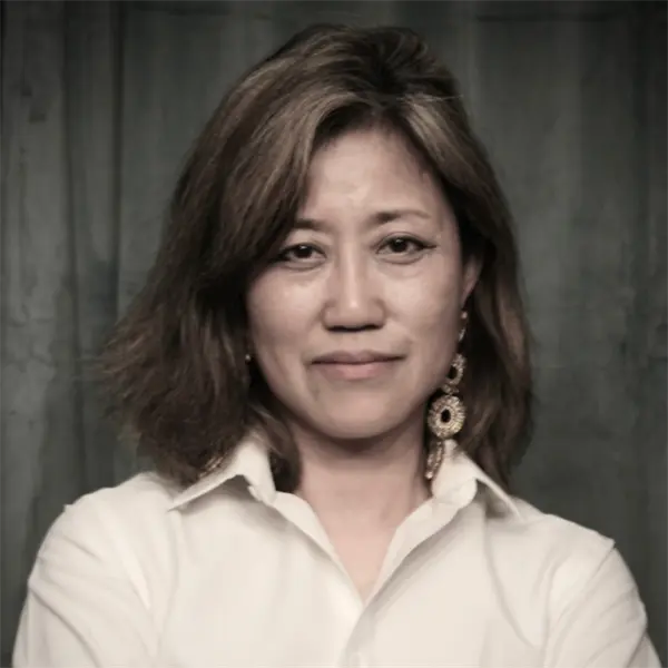 A woman with shoulder-length hair, wearing a white collared shirt and large earrings, stands in front of a dark background with her arms crossed.