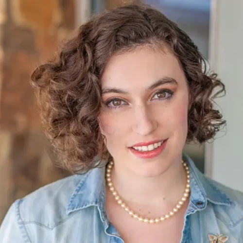A person with short, curly brown hair wears a pearl necklace and a light denim shirt, smiling at the camera. A blurred stone wall is in the background.