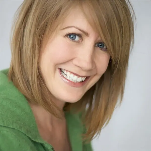 Woman with straight, shoulder-length blonde hair and blue eyes smiling, wearing a green top, in front of a light background.