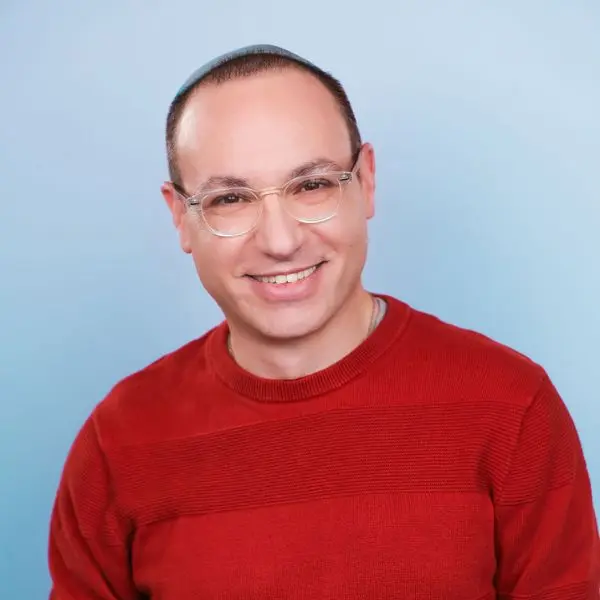 A person with short hair, wearing glasses and a red sweater, smiles at the camera against a plain light blue background.