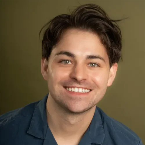 A young man with short brown hair and light eyes smiles at the camera, wearing a blue collared shirt against an olive-green background.