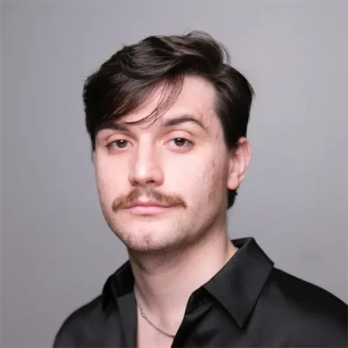 A young man with short dark hair, a mustache, and light stubble wears a black collared shirt and a thin necklace, standing against a plain light gray background.