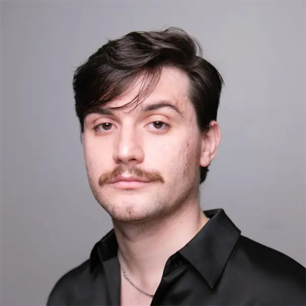 A young man with short dark hair, a mustache, and light stubble wears a black collared shirt and a thin necklace, standing against a plain light gray background.
