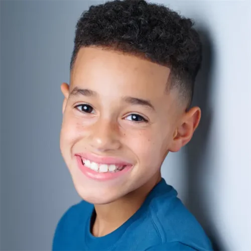 Smiling young boy with short curly hair wearing a blue shirt, standing against a light-colored wall.