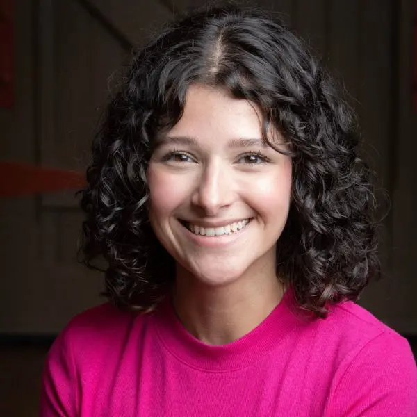 A woman with short curly dark hair wearing a bright pink top smiles at the camera against a dimly lit background.