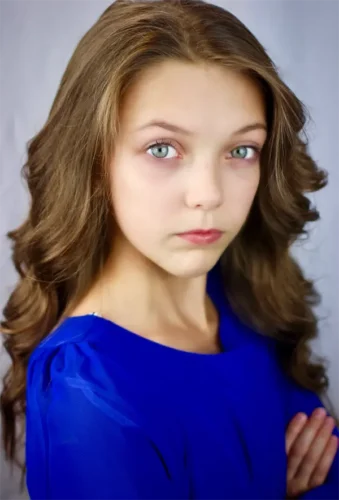 Girl with long brown hair and blue eyes wearing a royal blue top, standing with arms crossed and looking directly at the camera against a plain background.