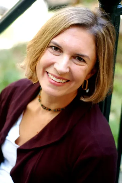A woman with short light brown hair, wearing a maroon jacket, white shirt, and beaded necklace, smiles while seated outdoors.