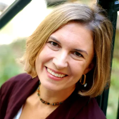 A woman with short light brown hair, wearing a maroon jacket, white shirt, and beaded necklace, smiles while seated outdoors.
