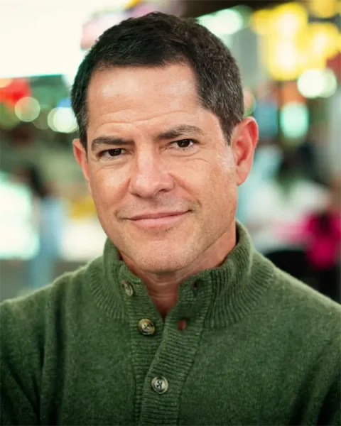 A man with short dark hair and a green button-up sweater stands indoors, looking directly at the camera with a neutral expression. The background is blurred.