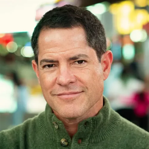 A man with short dark hair and a green button-up sweater stands indoors, looking directly at the camera with a neutral expression. The background is blurred.