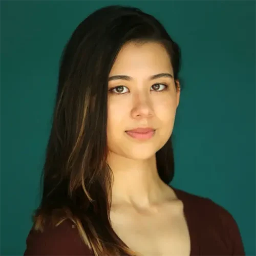 A young woman with long dark hair and a neutral expression looks at the camera, standing in front of a solid teal background.