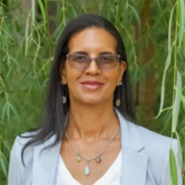 A woman with long dark hair, glasses, and a light-colored blazer stands outdoors in front of green foliage.