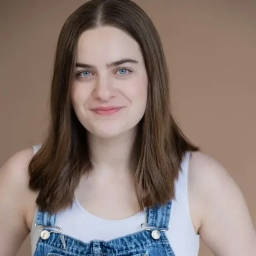 A person with straight brown hair wearing a white tank top and denim overalls stands against a plain light brown background, smiling slightly.