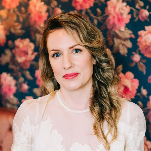 A woman with wavy blonde hair and a light-colored lace top poses in front of a dark floral wallpaper background.