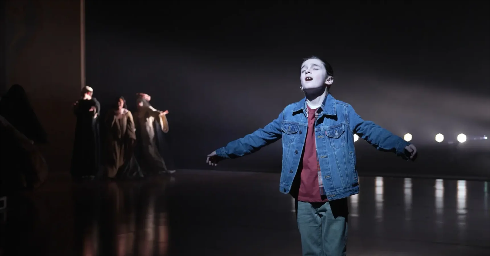 A young child in a denim jacket stands with arms outstretched under a spotlight, while a group of people in robes are seen in the background.