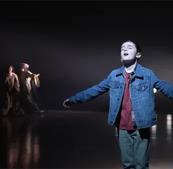 A young boy stands alone on stage with arms outstretched, singing, while several people in costumes stand in the dimly lit background.