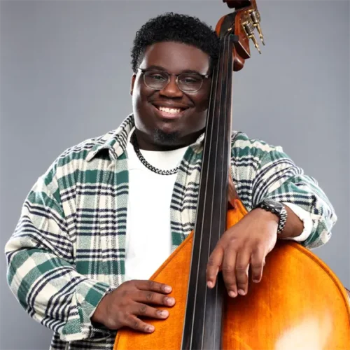 A man wearing glasses and a plaid shirt smiles while holding an upright bass against a plain gray background.
