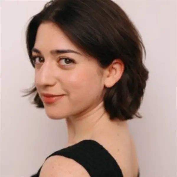 A woman with short dark hair and a black sleeveless top looks over her shoulder, smiling slightly against a plain light background.
