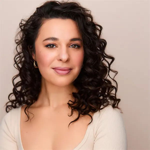 A woman with dark, curly hair wearing a cream-colored top and gold hoop earrings poses against a neutral background.