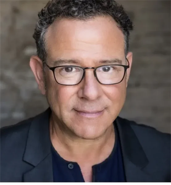 A middle-aged man with short curly hair, glasses, and a dark jacket looks at the camera against a blurred background.