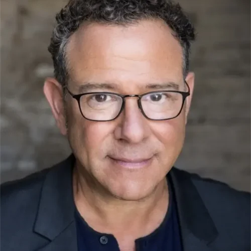 A middle-aged man with short curly hair, glasses, and a dark jacket looks at the camera against a blurred background.