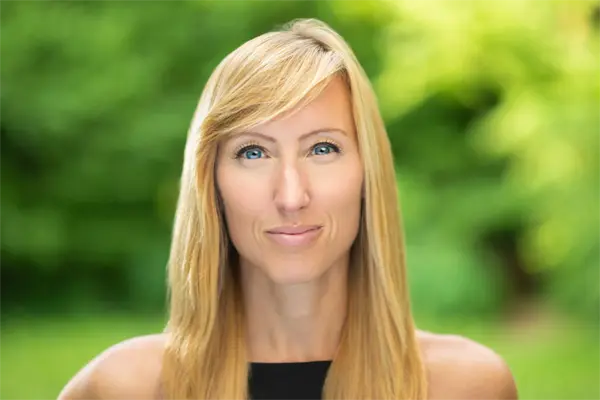 A woman with long blonde hair and blue eyes is standing outdoors, facing the camera, with a blurred green background.
