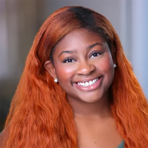 A young woman with long, wavy reddish-brown hair smiles at the camera, wearing a green top and stud earrings, with a blurred indoor background.