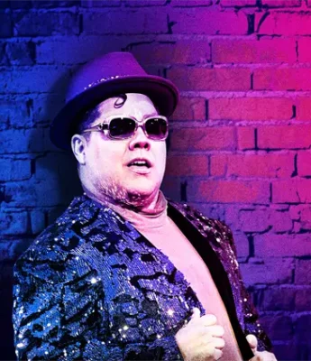 A person wearing sunglasses, a sequined jacket, and a purple hat poses in front of a brick wall lit with blue and pink lights.